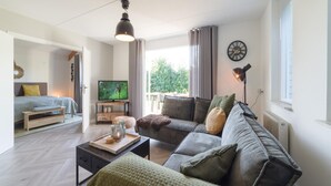 A modern living room with a sofa, coffee table, and a television. There is a bed in the adjacent room, and a window with curtains. - Holiday Home in Quiet Park with Garden (Wemeldinge)