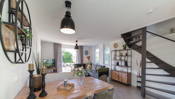 A spacious living area with a wooden dining table, comfortable seating, a staircase, and a bookshelf. - Holiday Home in Quiet Park with Garden (Wemeldinge)
