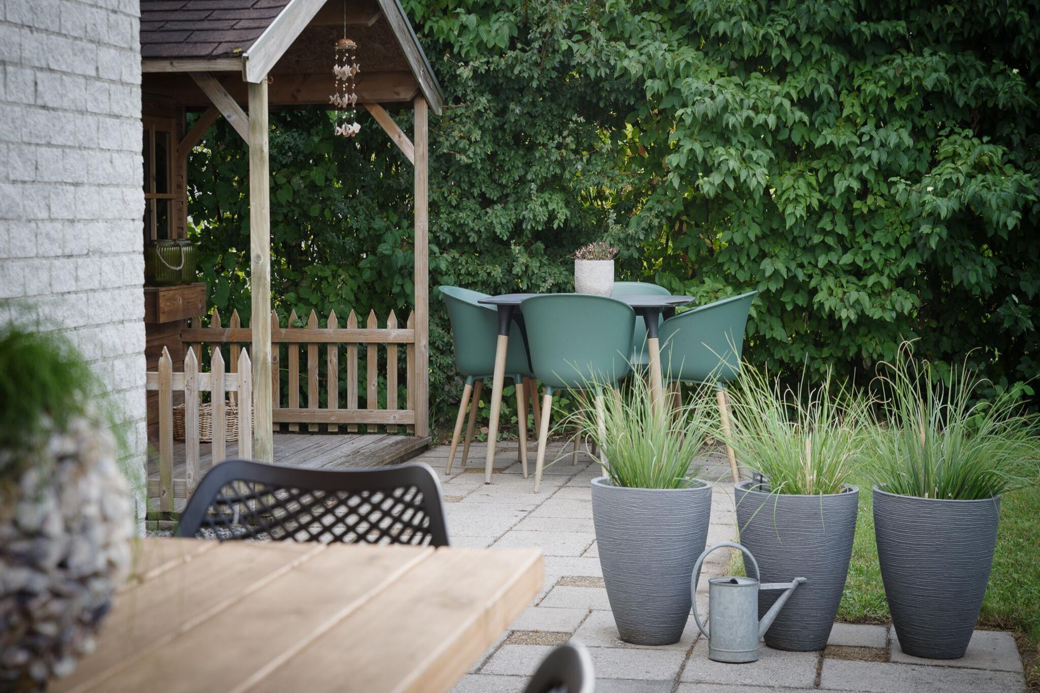An outdoor patio with a wooden table and chairs, surrounded by potted plants and a wooden fence.