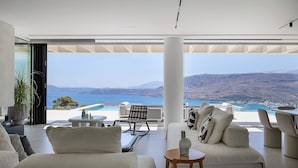 Ein modernes Wohnzimmer mit einem großen Glasfenster, das auf einen Balkon mit Blick auf eine malerische Landschaft aus Meer und Bergen führt.