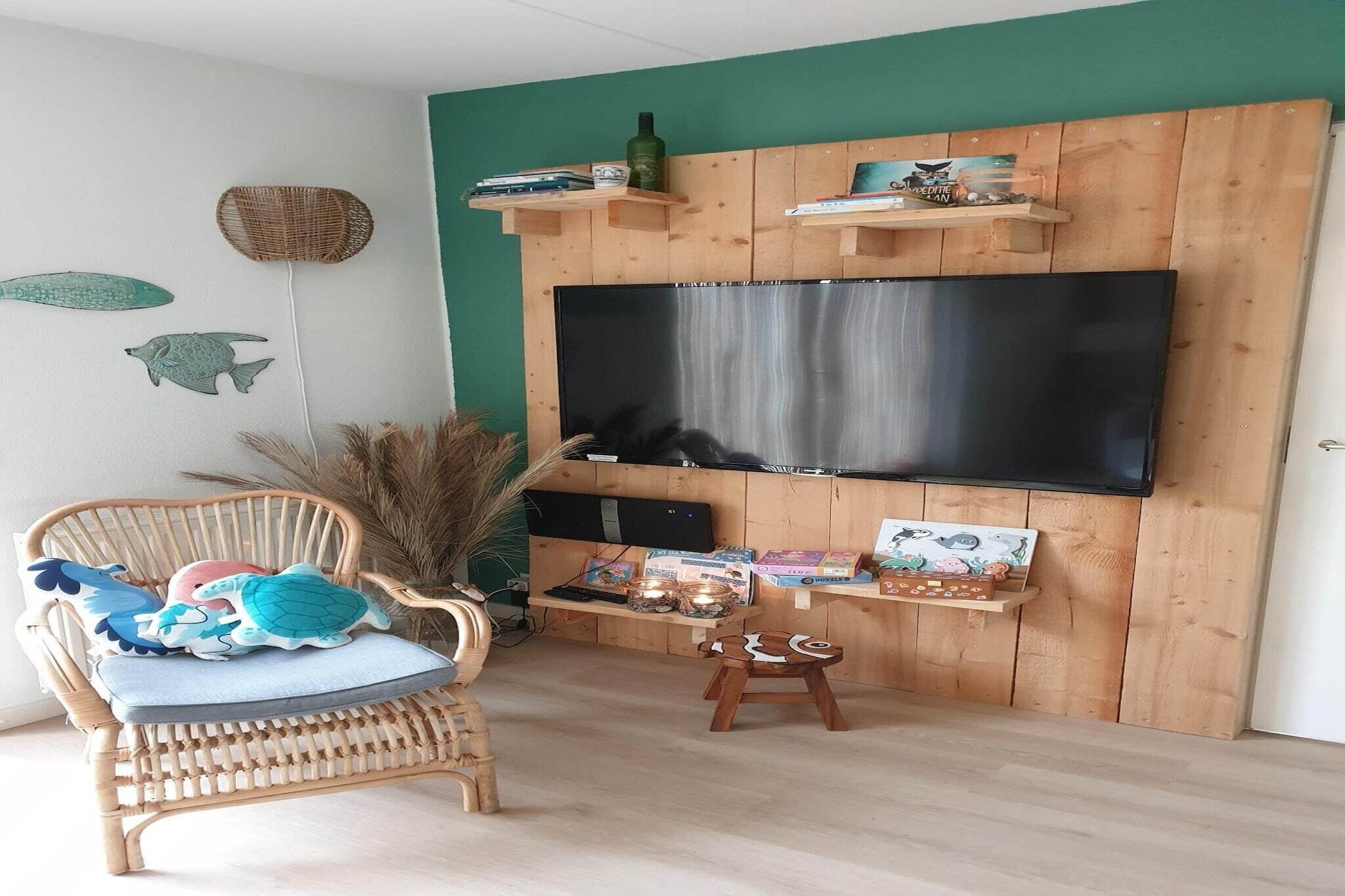 Una sala de estar con un televisor de pantalla plana grande montado en una pared de madera, un sillón de mimbre con cojín, un taburete pequeño y una estantería con adornos.
