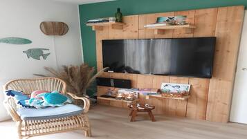 Una sala de estar con un televisor de pantalla plana grande montado en una pared de madera, un sillón de mimbre con cojín, un taburete pequeño y una estantería con adornos.