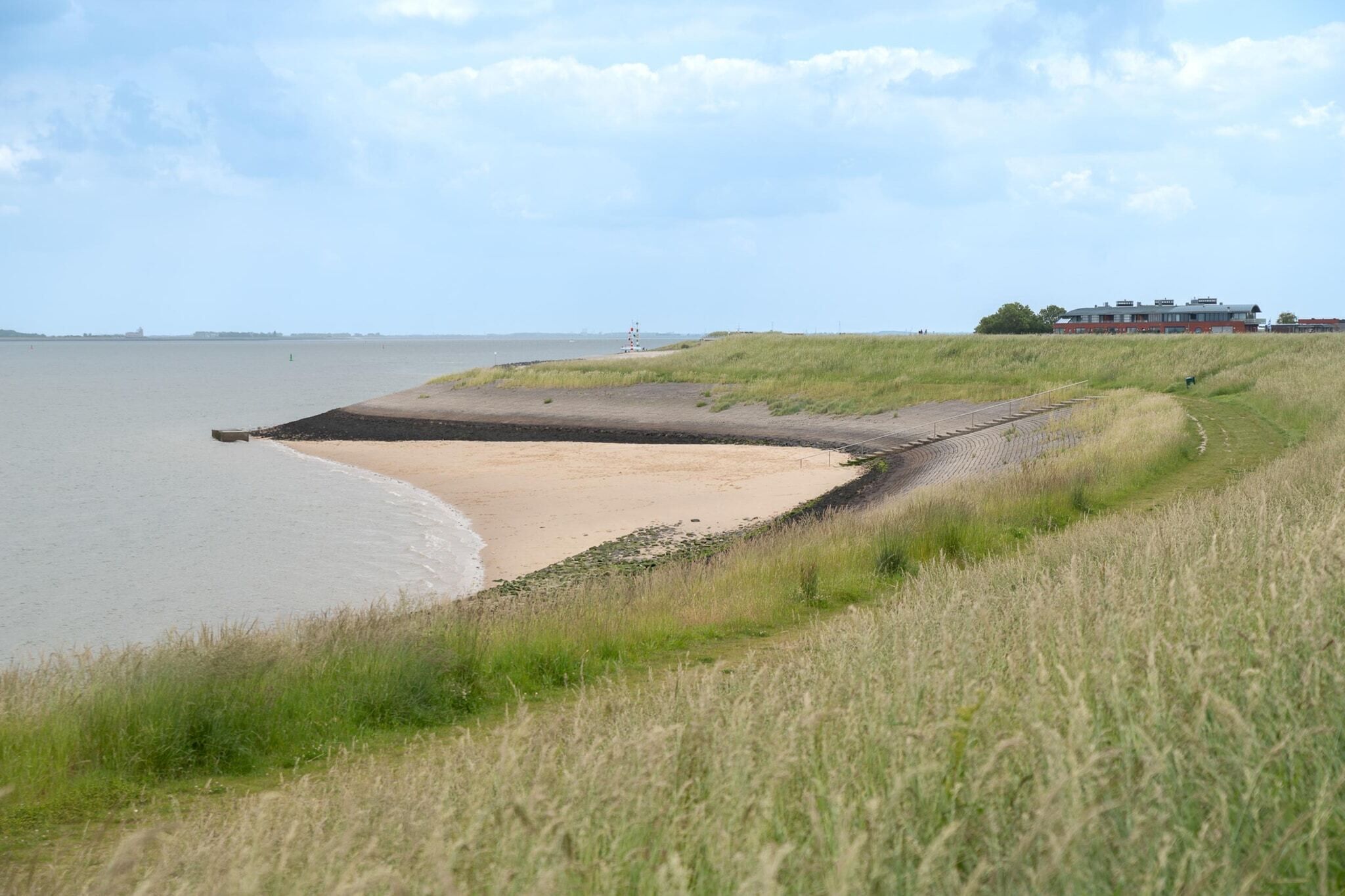Un paisaje costero con una playa de arena, dunas cubiertas de hierba y un edificio al fondo.