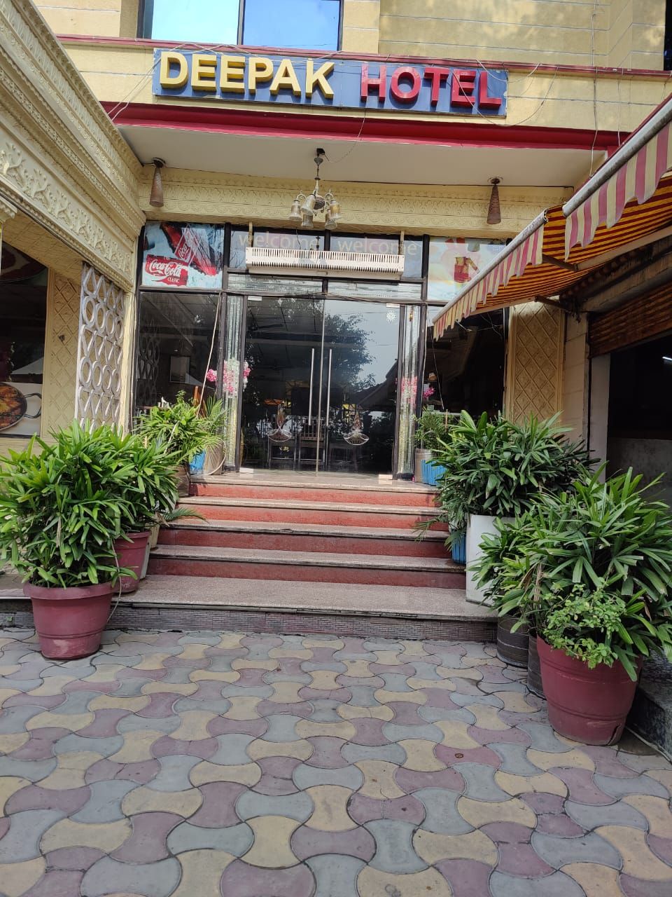 The entrance to Deepak Hotel with a Coca-Cola sign, potted plants, and a tiled pavement.