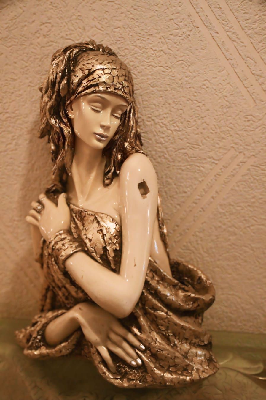 A statue of a woman in a dress with a textured pattern, placed on a surface against a textured wall.