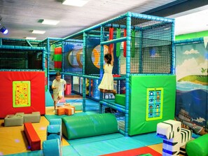 An indoor play area with netted climbing structures, padded flooring, and colorful play mats.