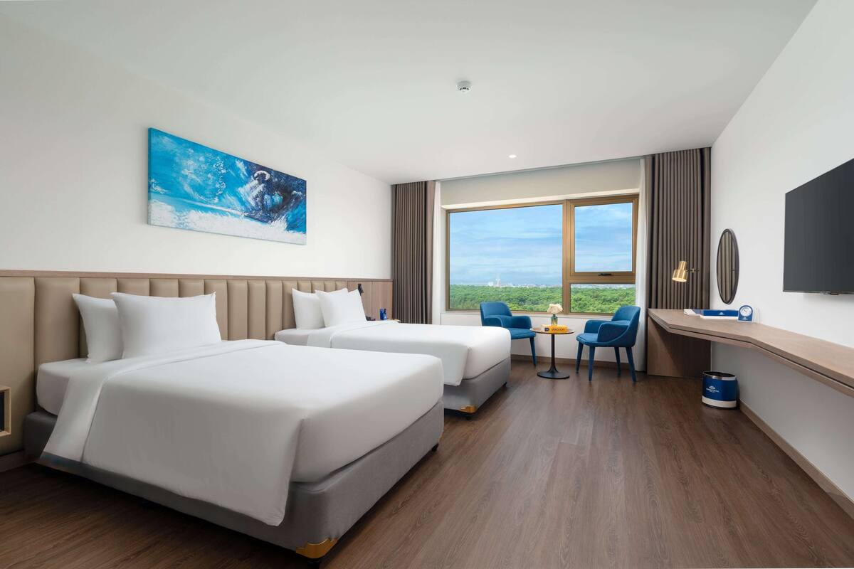 Habitación de hotel con dos camas, un escritorio, una silla, un ventanal con vistas y un cuadro en la pared.
