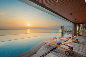 Une piscine à débordement moderne, dotée de chaises longues en bois et d’une balustrade en verre, offrant une vue sur la mer au coucher du soleil.