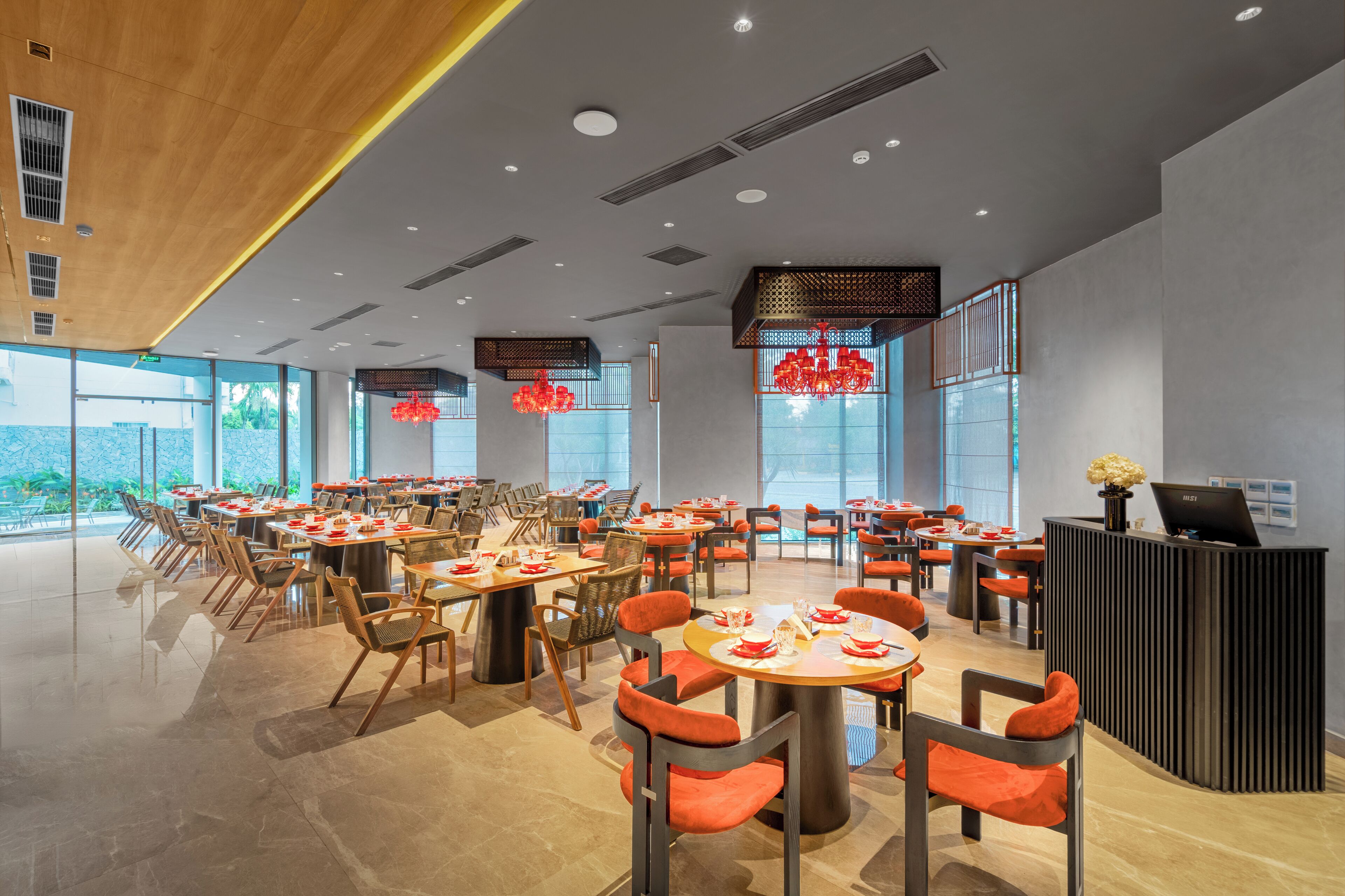 A modern restaurant with multiple dining tables and chairs, a reception desk, and large windows offering a view of the outdoors.