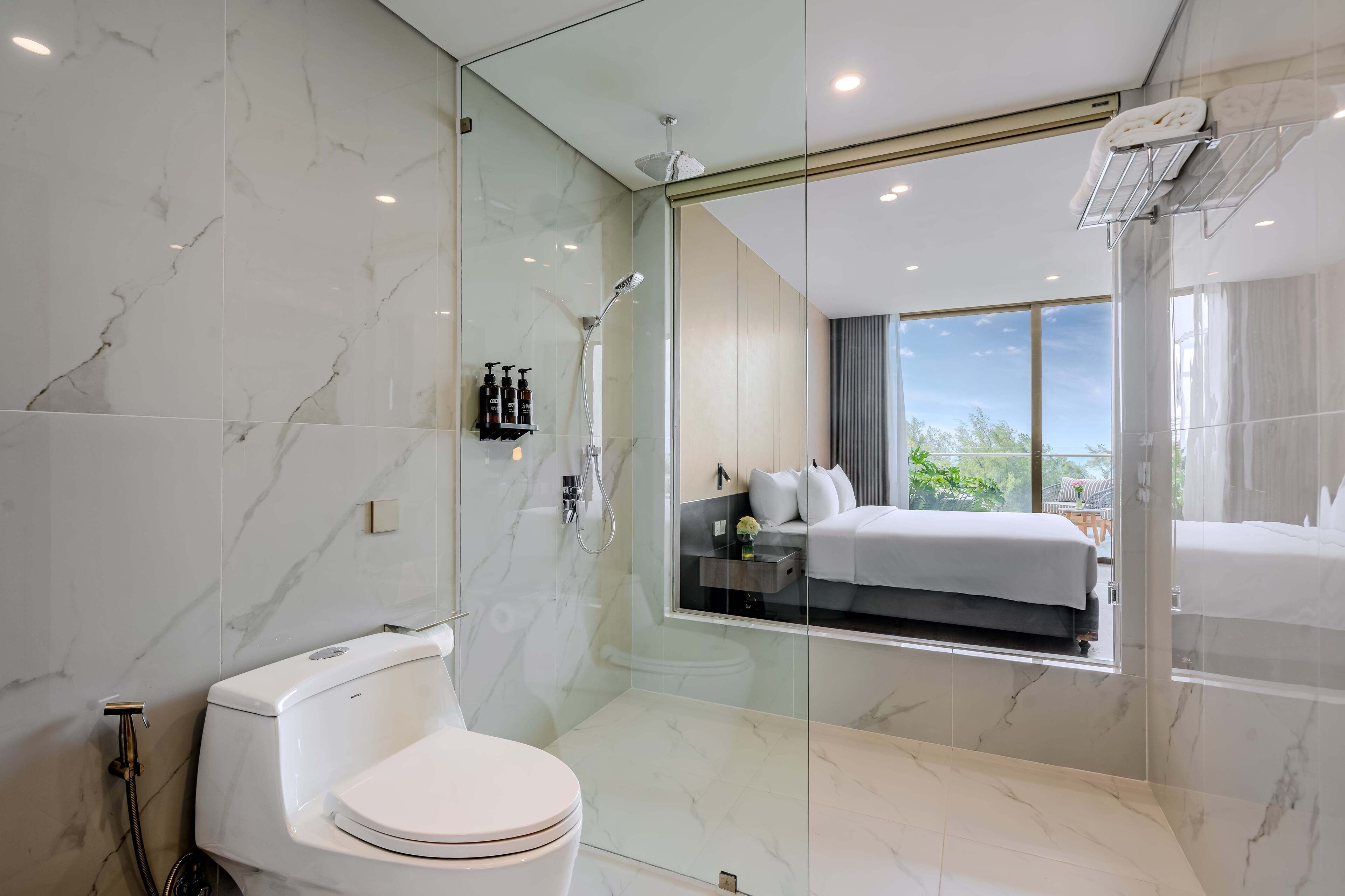 Un baño moderno con ducha envuelta en vidrio, inodoro y un espejo grande que refleja un dormitorio con cama y ventana.