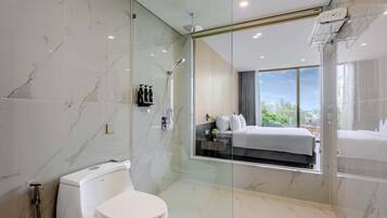 Un baño moderno con ducha envuelta en vidrio, inodoro y un espejo grande que refleja un dormitorio con cama y ventana.