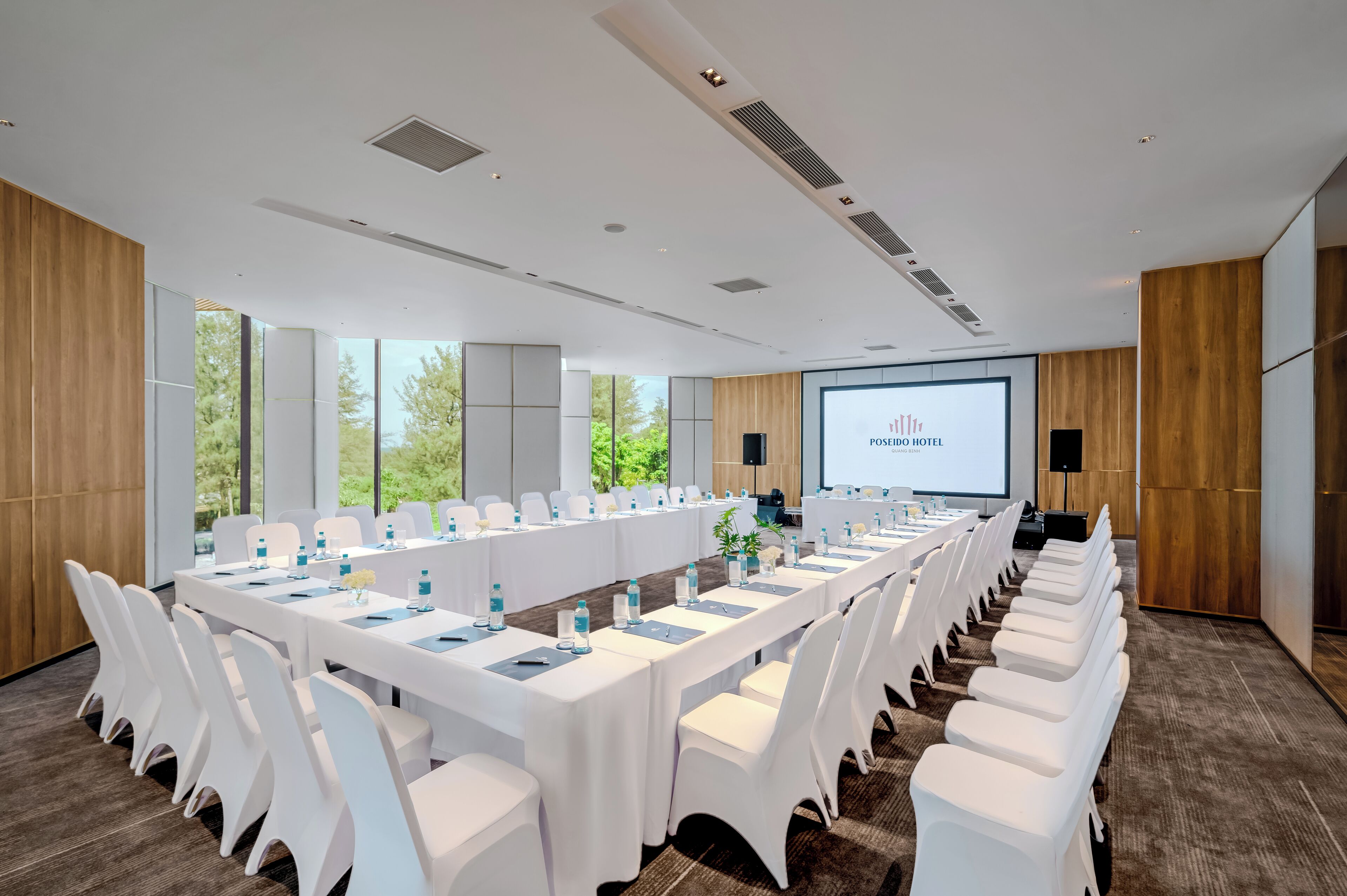 A conference room with a long table, white chairs, and a projector screen displaying "POSIDO HOTEL".