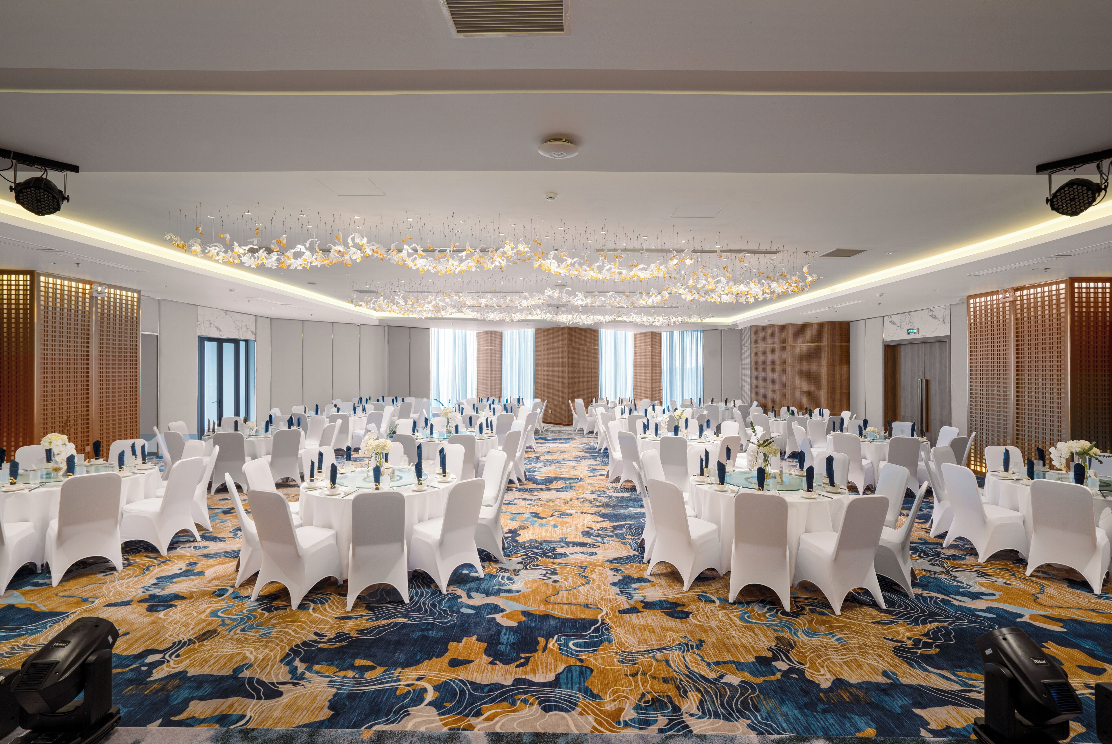 A large conference room with rows of white chairs, tables with blue centerpieces, and a patterned carpet.