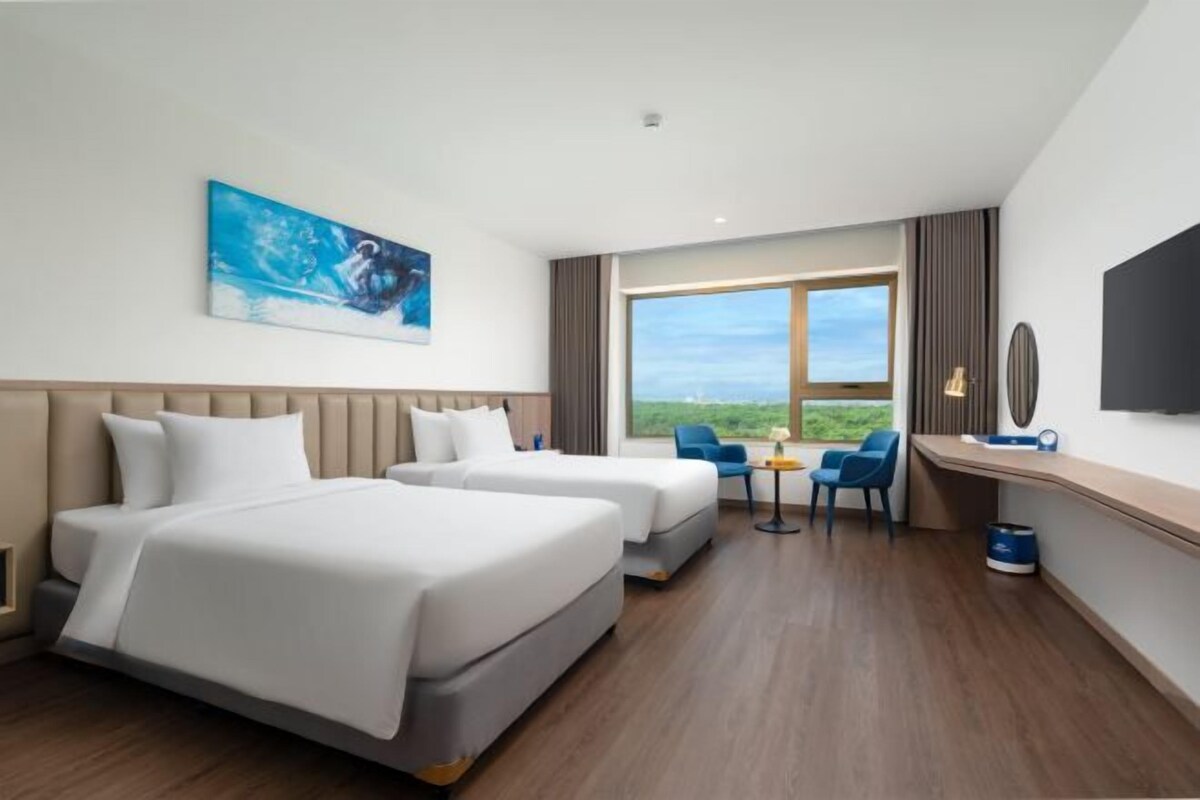 A hotel room with two beds, a desk, a chair, and a large window offering a view.