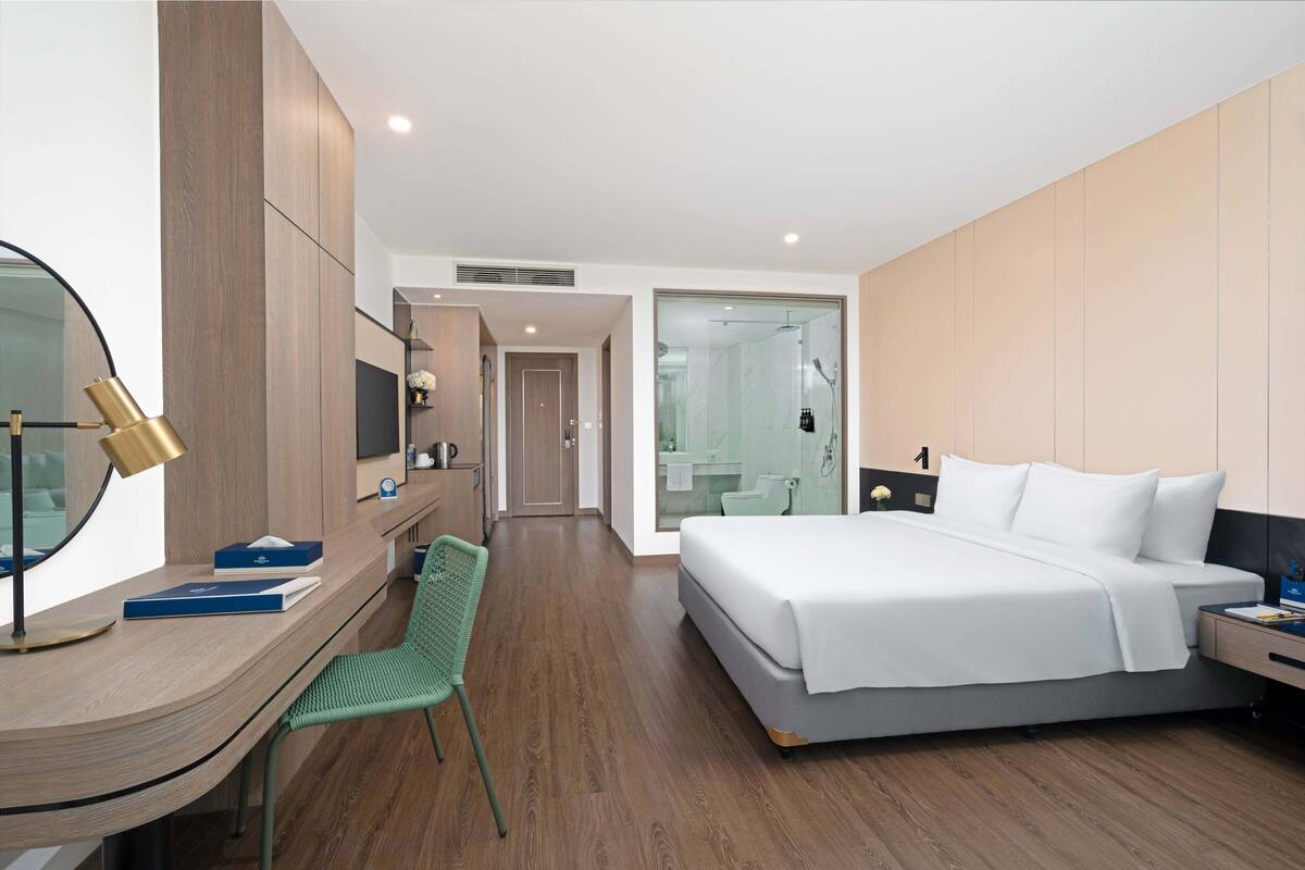 Una habitación de hotel moderna con un escritorio de madera, una silla verde, una cama grande con ropa blanca, una mesita de noche y un espejo.