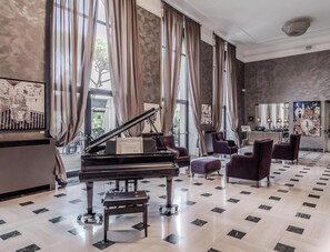 A grand piano in a spacious room with a patterned floor, large windows with curtains, and a seating area with chairs. - HOTIDAY Room Collection - Roma Cornelia (Rome)
