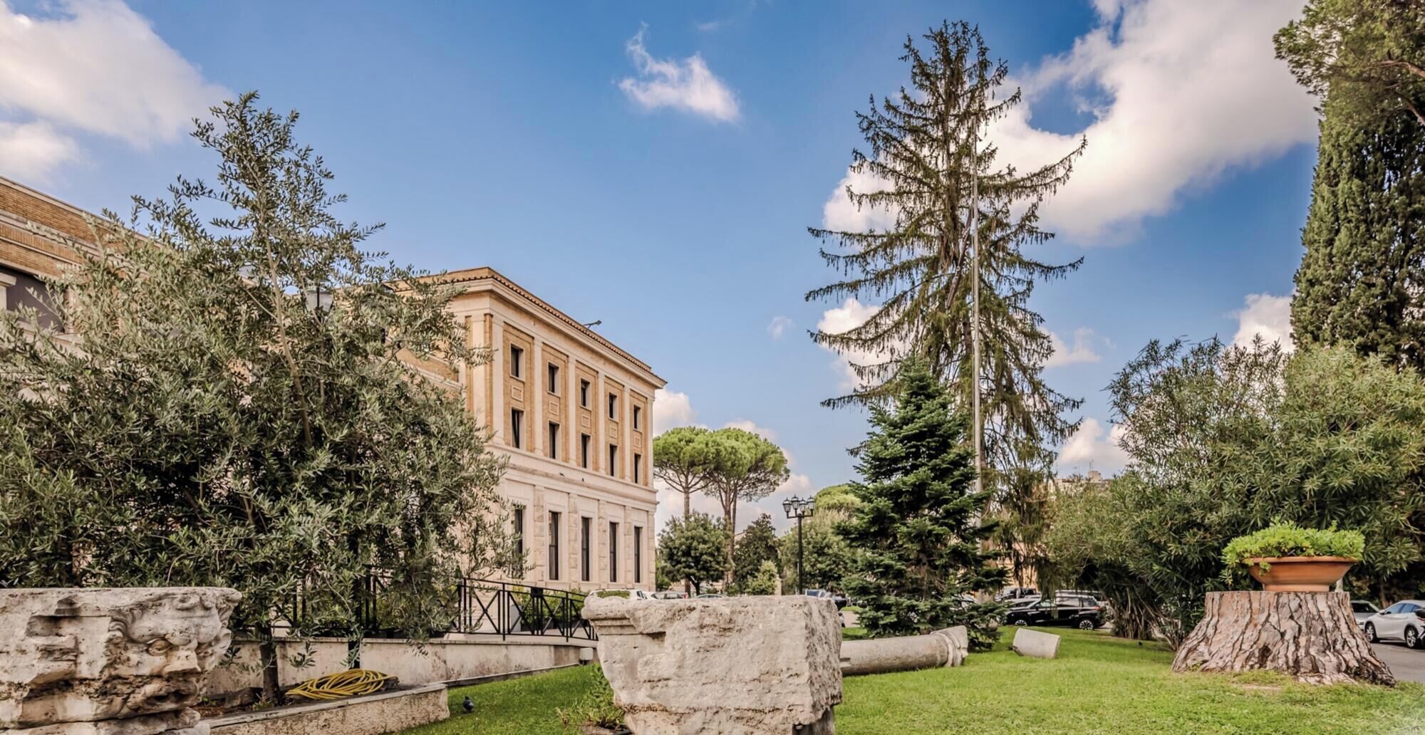 A courtyard with a classical building, stone structures, and a large tree.