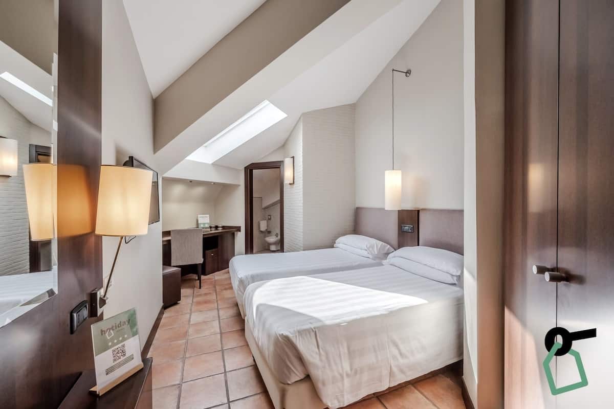 A hotel room with two beds, a desk, a bathroom, and a skylight.