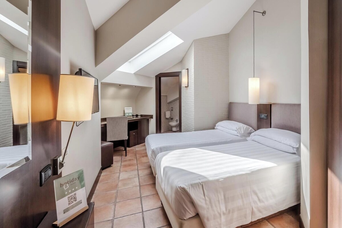 A hotel room with two beds, a desk, a bathroom, and a skylight.