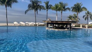 En klarblå pool med en liten paviljong omgiven av palmer och strandparasoller.