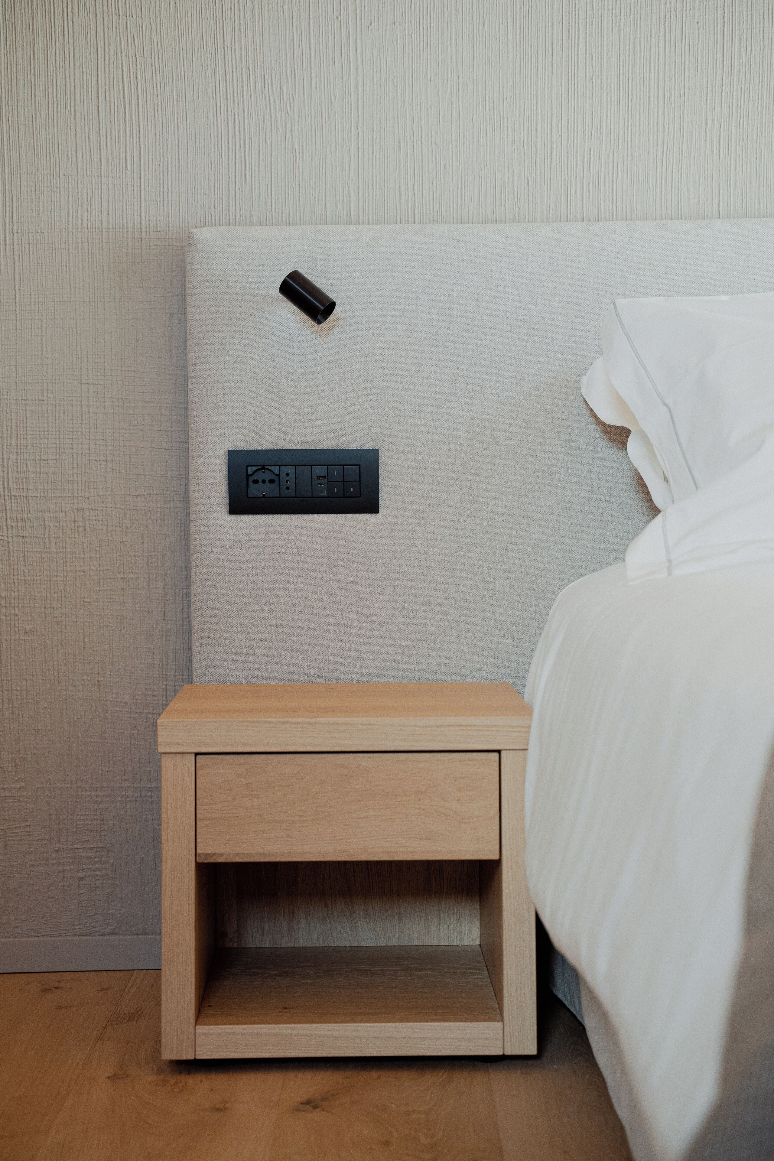 A minimalist bedroom with a wooden nightstand, a power outlet, and a wall-mounted control panel.