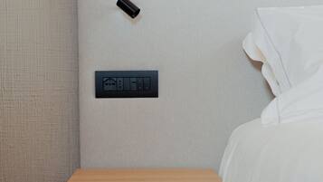 A minimalist bedroom with a wooden nightstand, a power outlet, and a wall-mounted control panel.