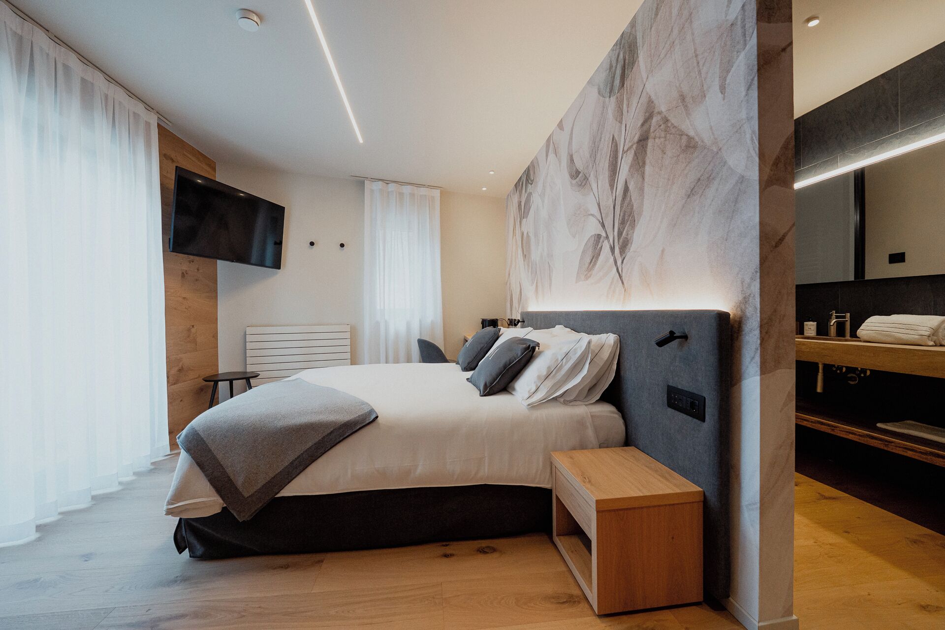 A modern bedroom with a large bed, bedside tables, a TV, and a bathroom with a mirror and sink.