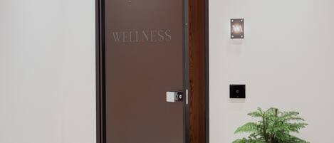 Een deur met daarop de tekst 'WELLNESS', een kleine plant in een pot en een wandlamp.