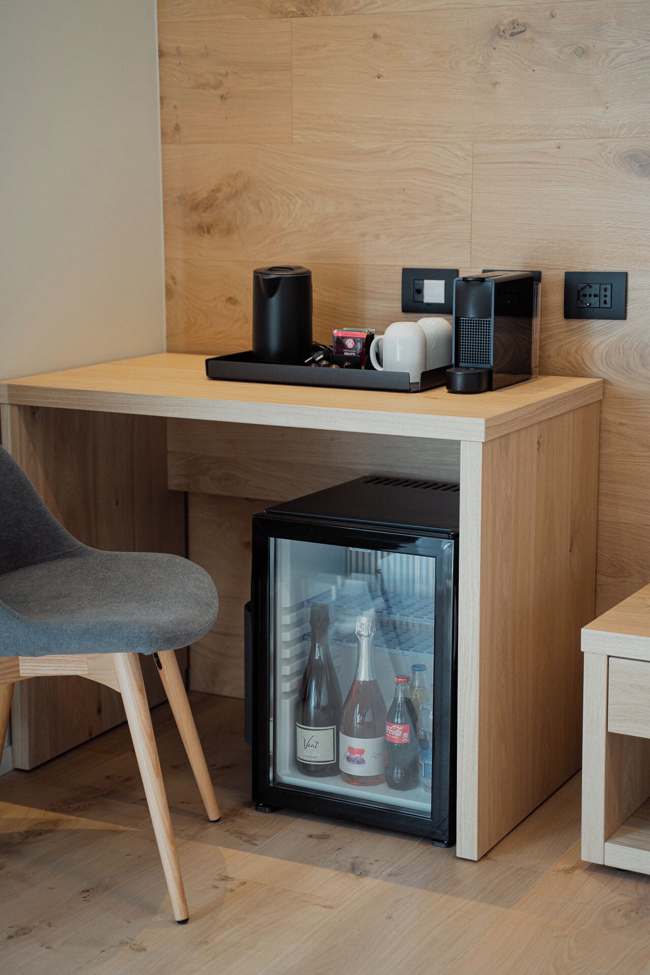 A compact kitchenette with a refrigerator containing beverages, a coffee maker, and a kettle on a wooden countertop.