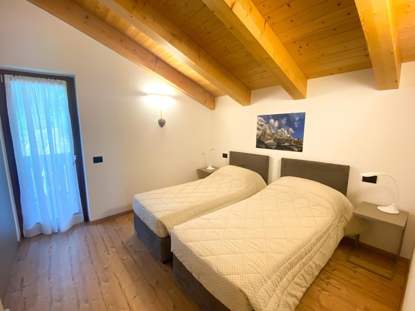 A bedroom with two beds, wooden beams on the ceiling, a nightstand with a lamp, and a window with curtains.
