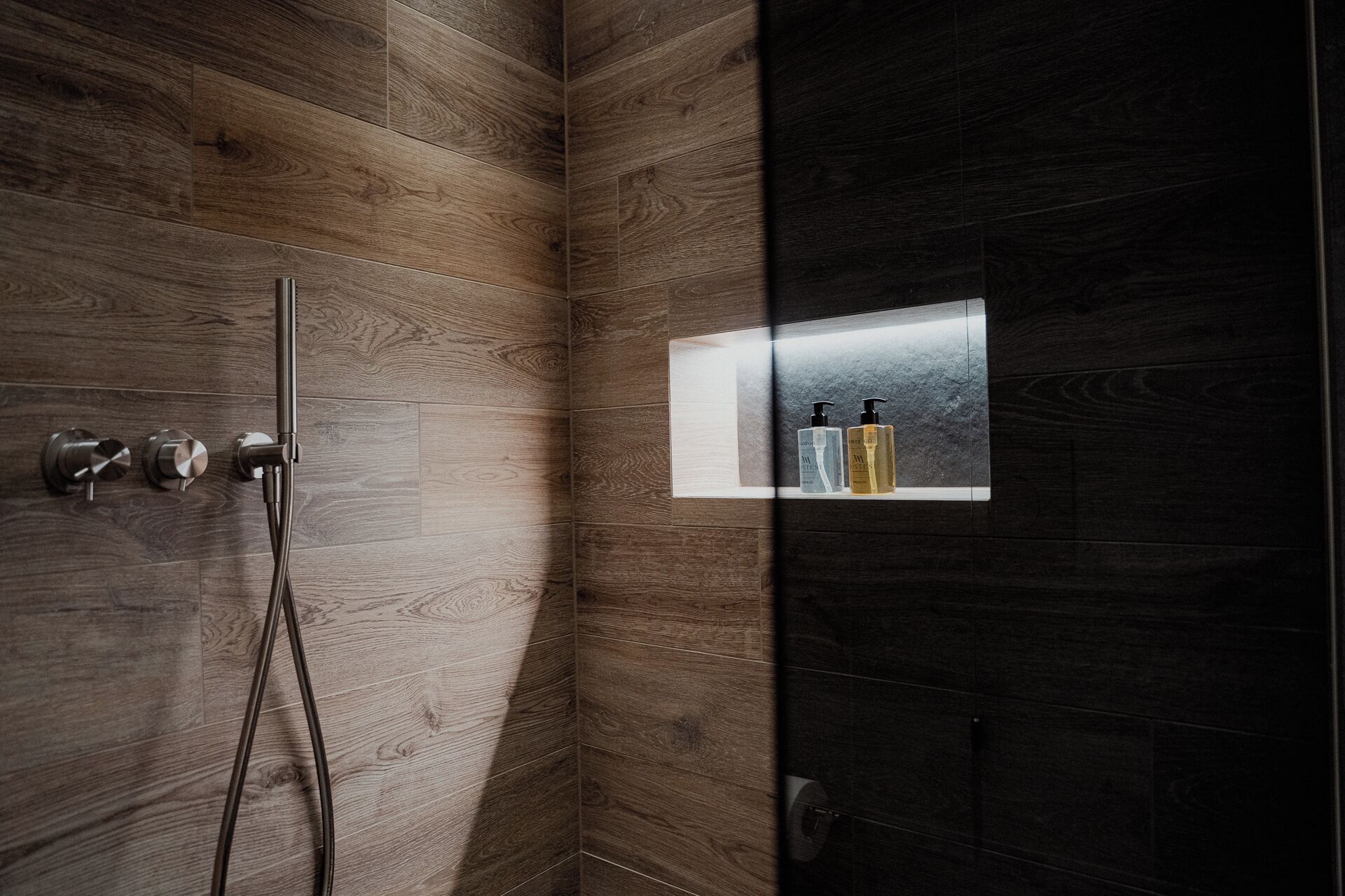 A shower area with wooden walls, a glass-enclosed shelf with toiletries, and a handheld showerhead.