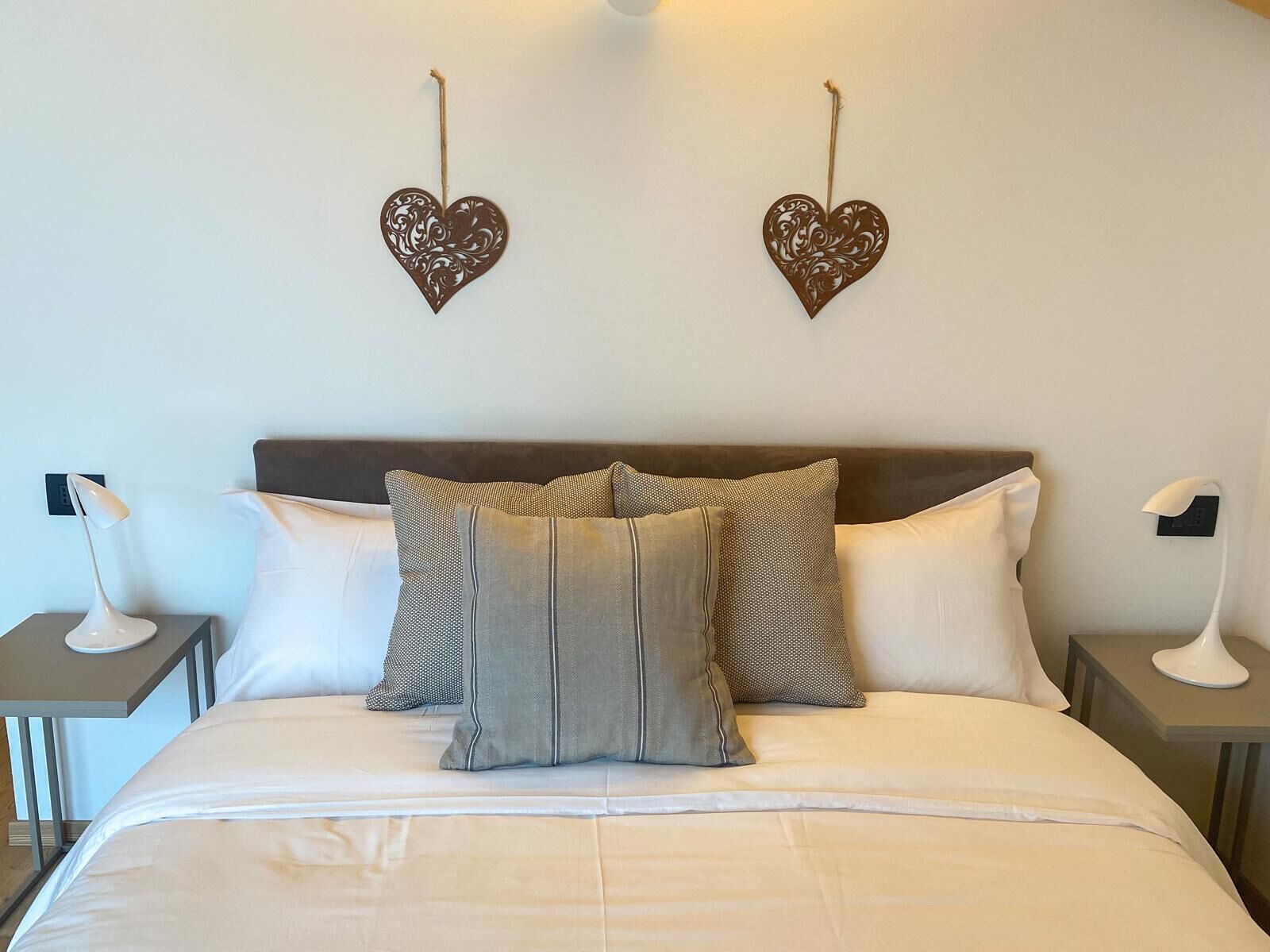 Una cama bien hecha con edredón blanco, dos almohadas y un cabecero. Hay dos mesitas de noche con lámparas y un adorno con forma de corazón en la pared.
