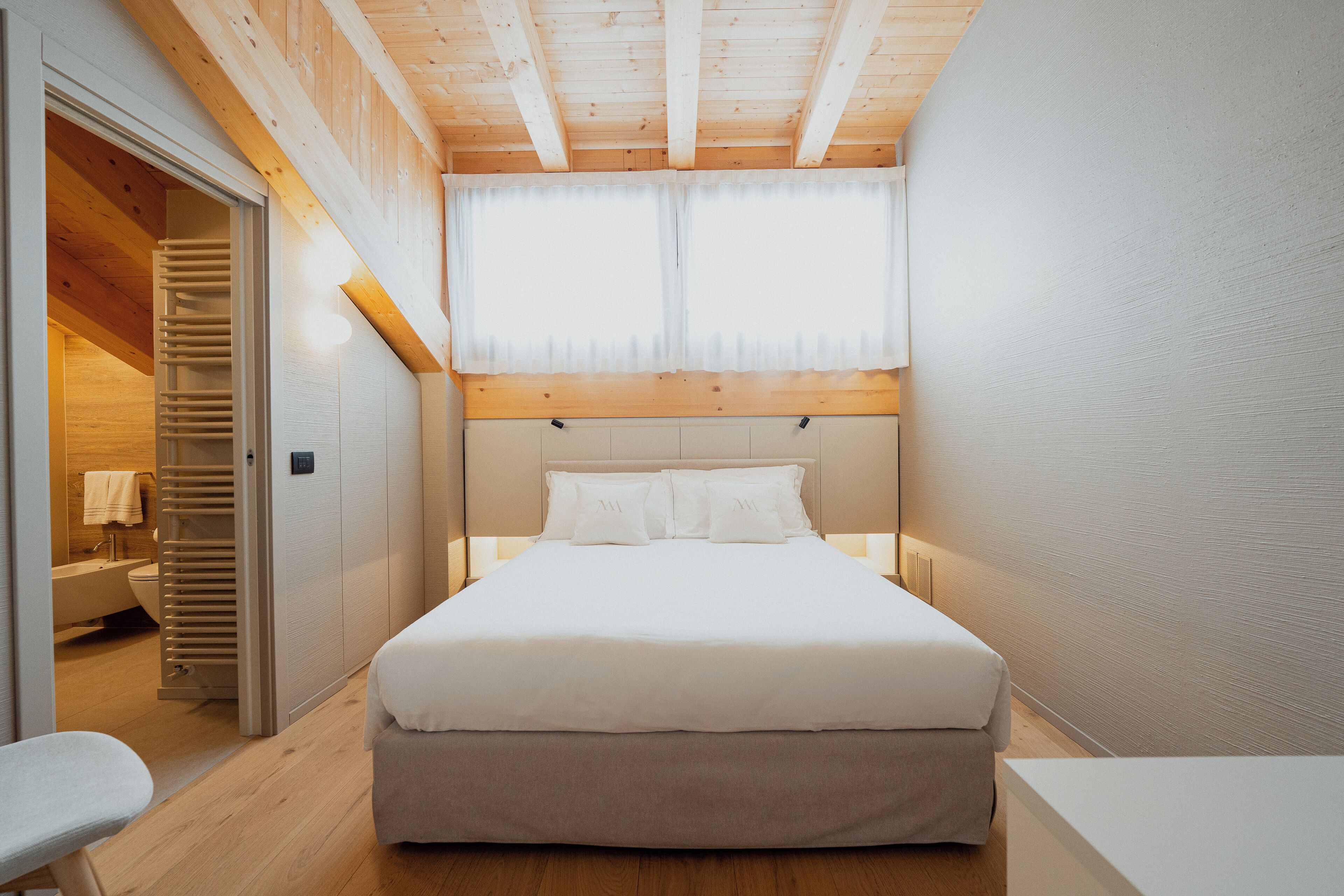 A bedroom with a wooden ceiling, a large bed with white bedding, a bedside table, and a window with sheer curtains.