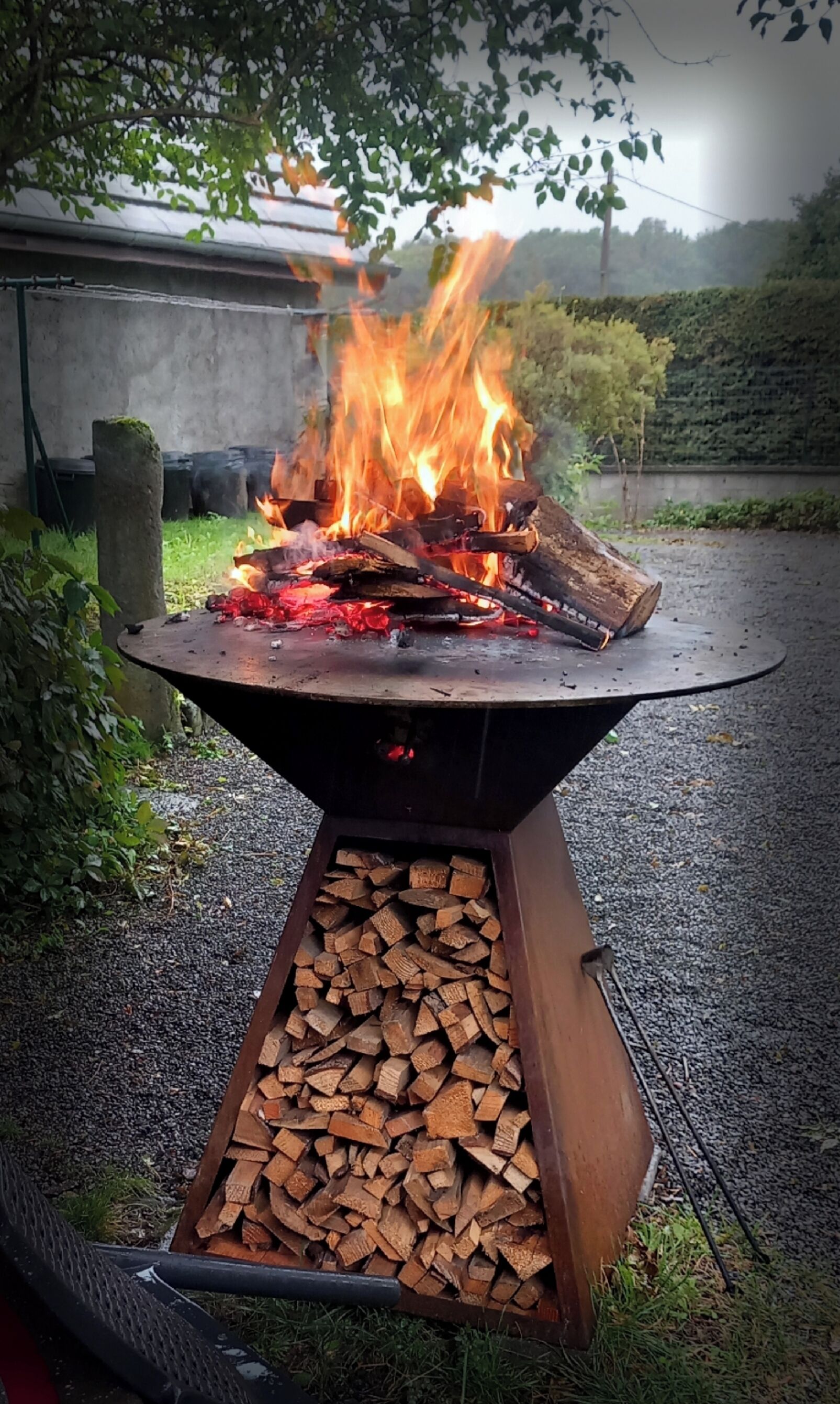 A metal fire pit with burning logs and flames, surrounded by gravel and greenery.