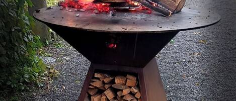 A metal fire pit with burning logs and flames, surrounded by gravel and greenery.