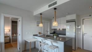 Una cocina moderna con una isla central, taburetes blancos tipo barra, iluminación colgante y vistas al dormitorio contiguo.