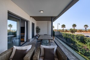 A balcony with a dining table and chairs, a sofa, and a glass railing overlooking a beach with palm trees.