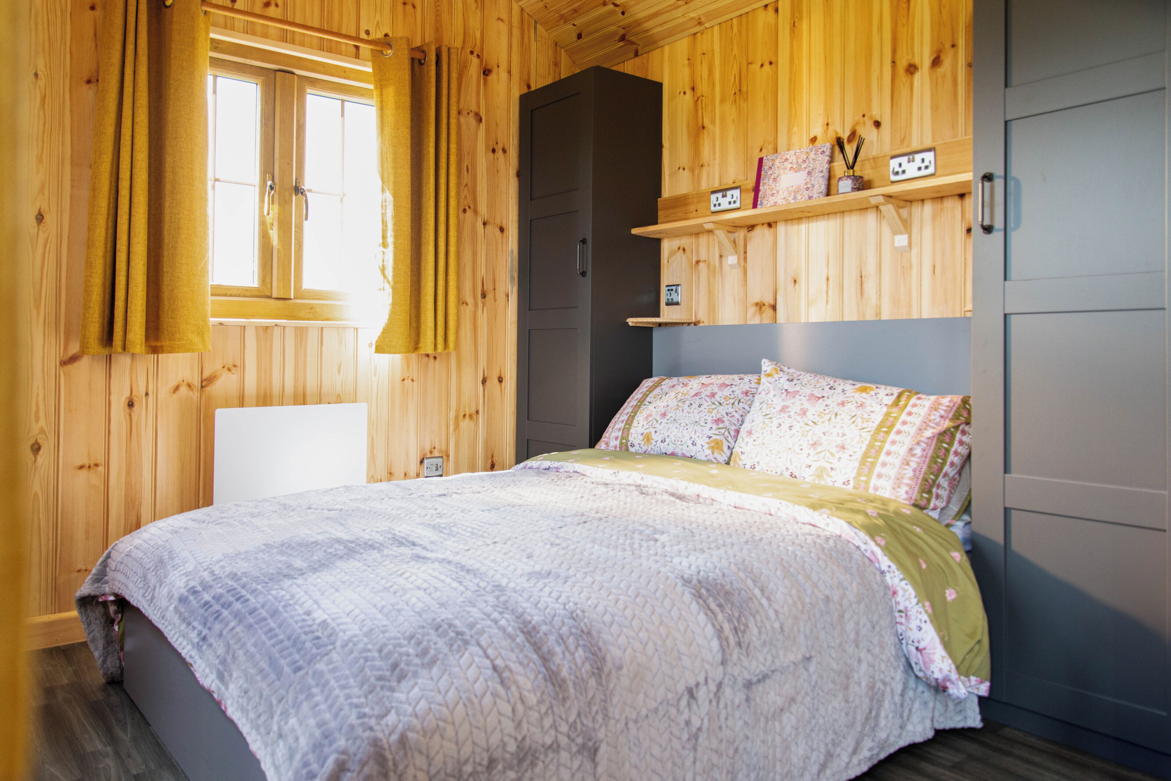 Una stanza in legno con un letto, una finestra con tende e uno scaffale con oggetti.