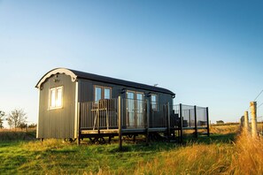 Une petite maison à un seul étage, dotée d’un porche et d’une terrasse entourée d’une clôture, au milieu d’une herbe haute et sous un ciel dégagé.