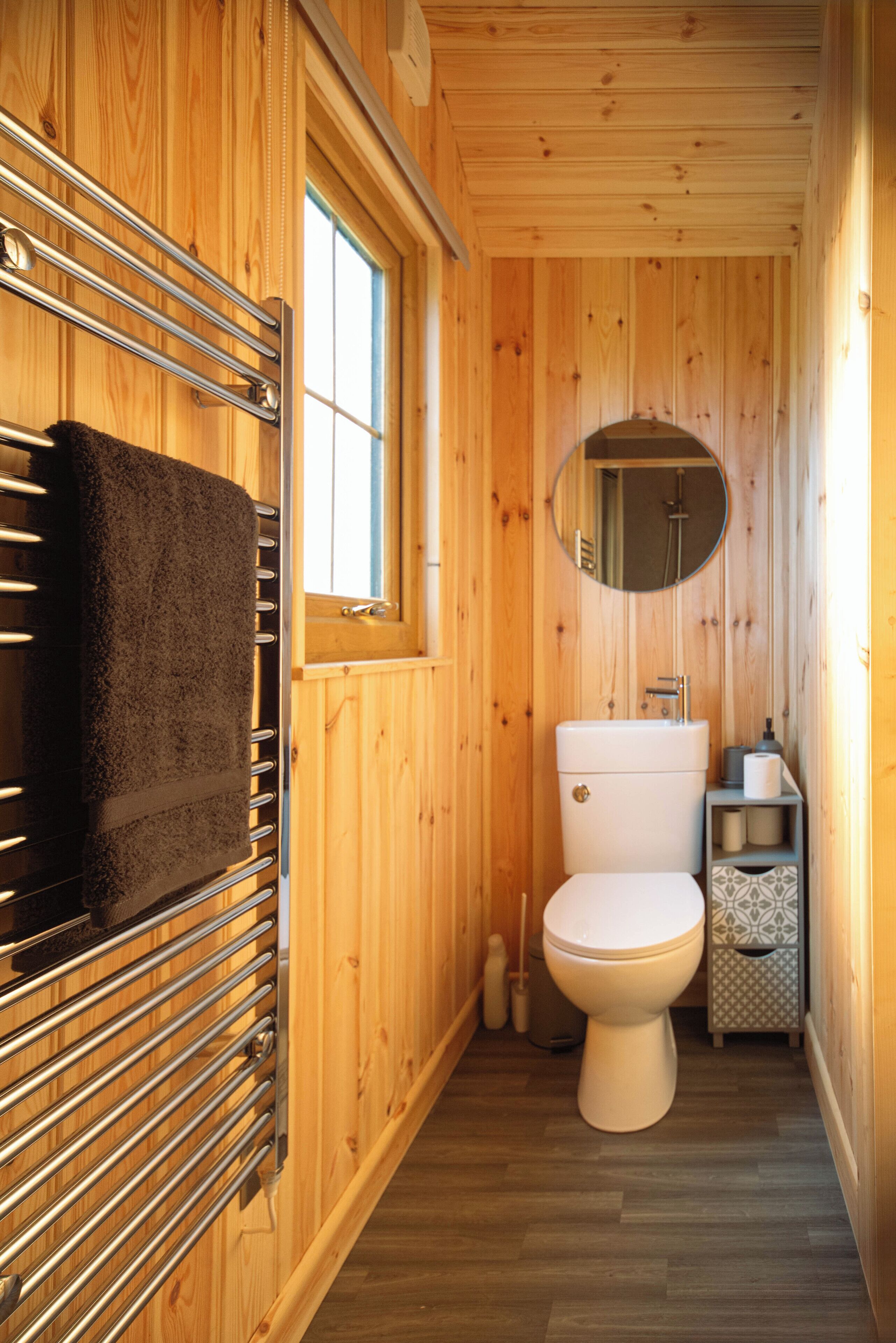 Une salle de bain aux murs lambrissés, avec des toilettes blanches, un porte-serviettes sur lequel est posée une serviette noire, un miroir rond et une fenêtre.