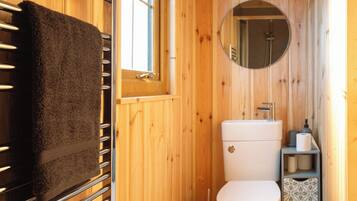 Une salle de bain aux murs lambrissés, avec des toilettes blanches, un porte-serviettes sur lequel est posée une serviette noire, un miroir rond et une fenêtre.