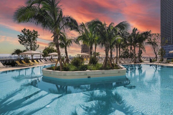 A swimming pool with lounge chairs and palm trees, surrounded by a high-rise building.