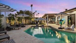 A pool area with lounge chairs and umbrellas, surrounded by palm trees and a modern building. - Bella Palma: Elegance & Style w/Putt, Pool & Spa! (Scottsdale)