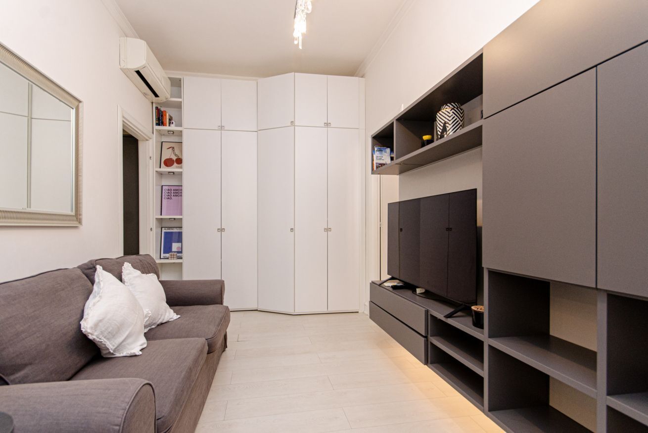 A modern living room with a grey sofa, white cushions, a flat-screen TV, and built-in shelving.