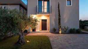A modern two-story house with a balcony, a well-lit entrance, and a landscaped garden.