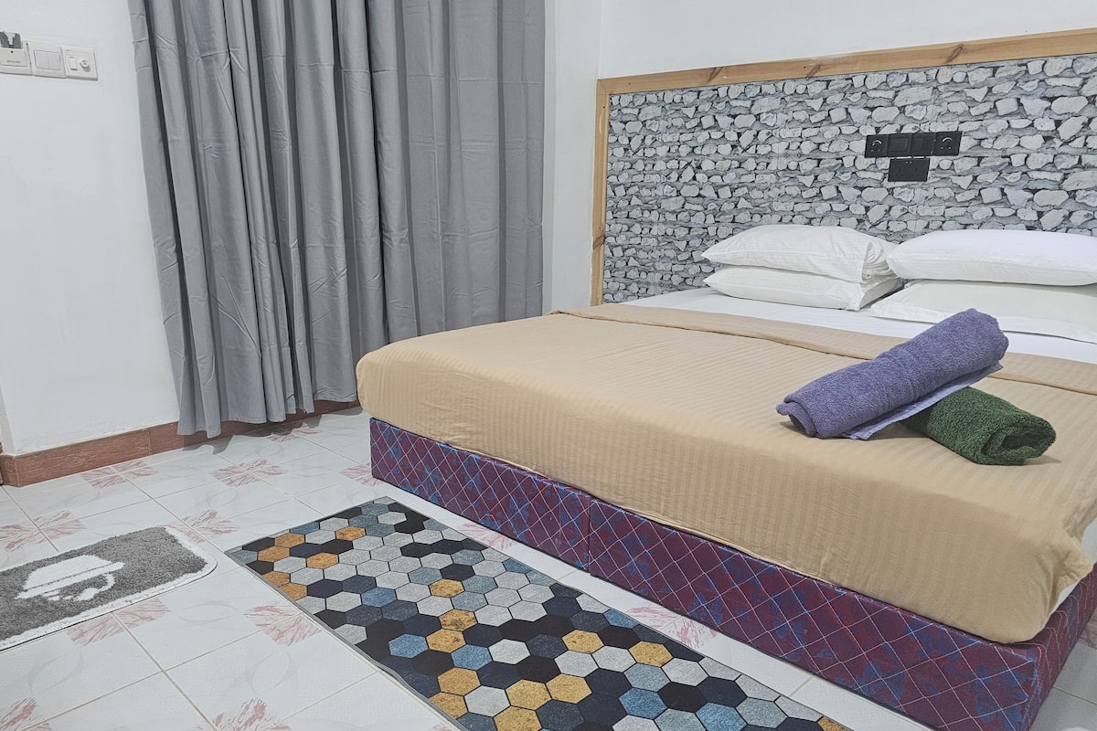 A bedroom with a bed, curtains, a headboard with a stone-like texture, and a patterned rug.