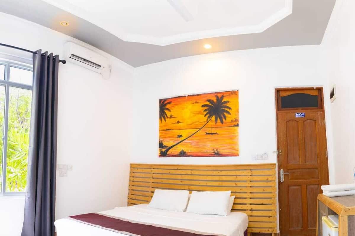 A bedroom with a bed, a painting of a sunset, a wooden door, and an air conditioning unit.