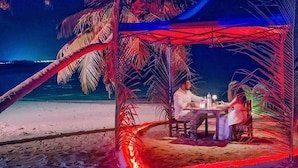 Un área de comedor frente a la playa, con mesa y sillas, palmeras y vista al océano.