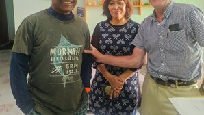 Tres personas de pie en un interior con un cuadro de playa en la pared y un estante de madera con diversos objetos.