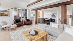A spacious living room with a high ceiling, wooden beams, a large sofa, leather armchairs, and a dining area with blue chairs. - Ski-In/Ski-Out via Pony Express Lift (Big Sky)